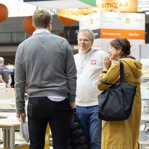 Ein Fachberater von ASS Einrichtungssysteme unterhält sich auf der Didacta neben einer Tischgruppe mit zwei Messebesuchern